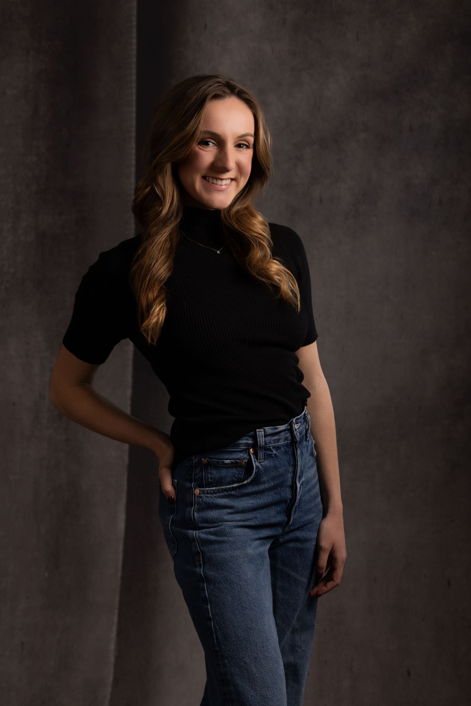 Young woman in black turtleneck top and jeans smiling confidently against a dark studio backdrop.