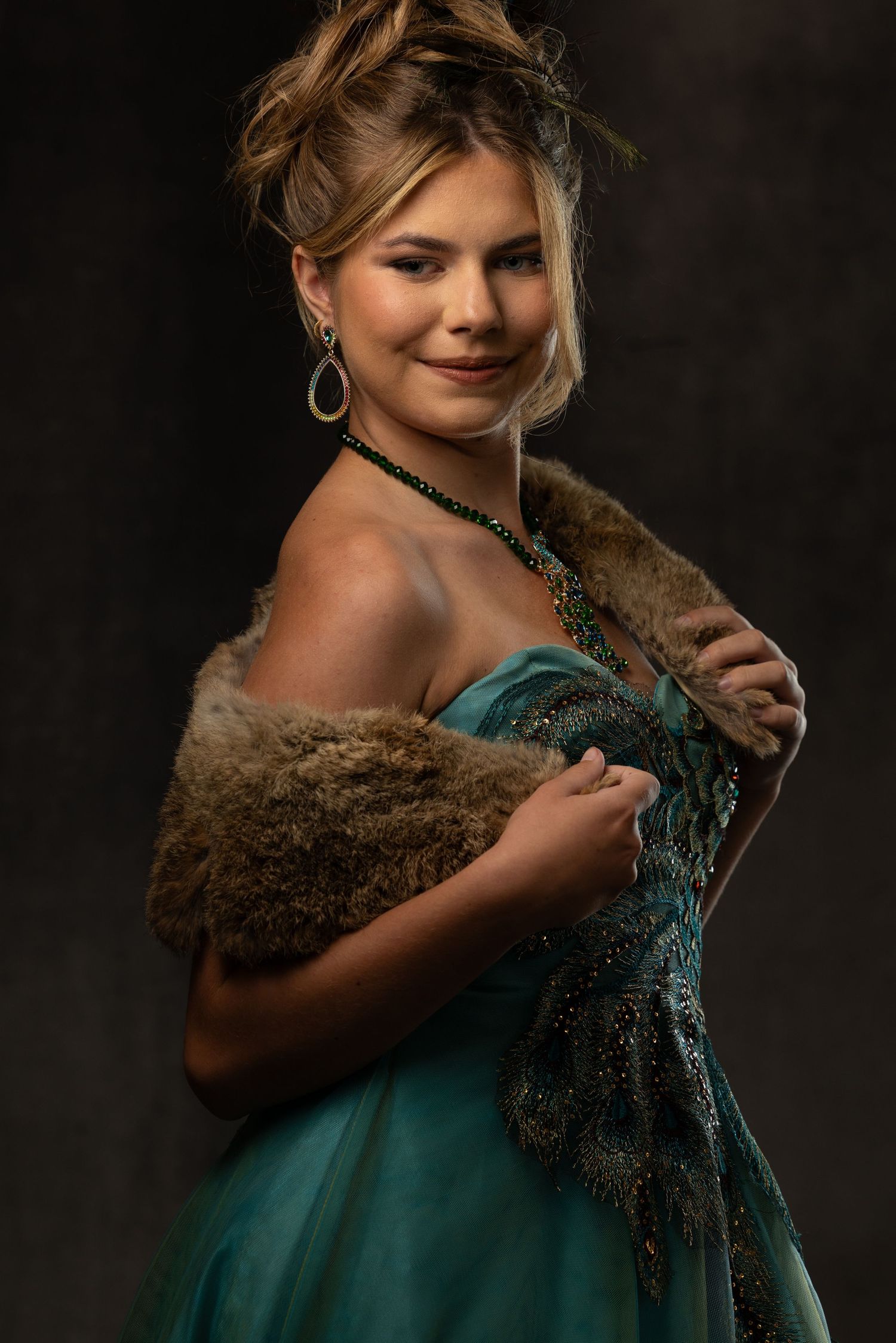 Young blonde woman in teal off-shoulder gown with fur stole and green jewelry poses elegantly against dark background.