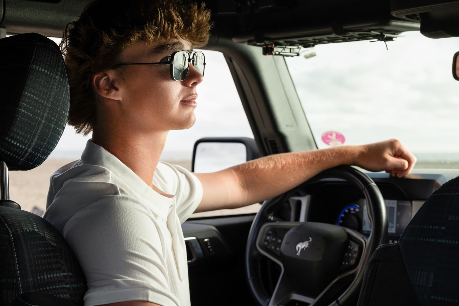 Young man with sunglasses driving a dark SUV, hand on steering wheel, looking ahead confidently.