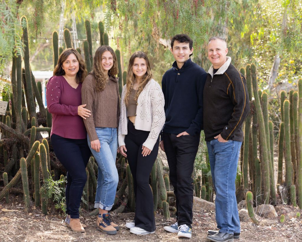 Family of five smiling together outdoors near tall cacti in a desert garden setting.