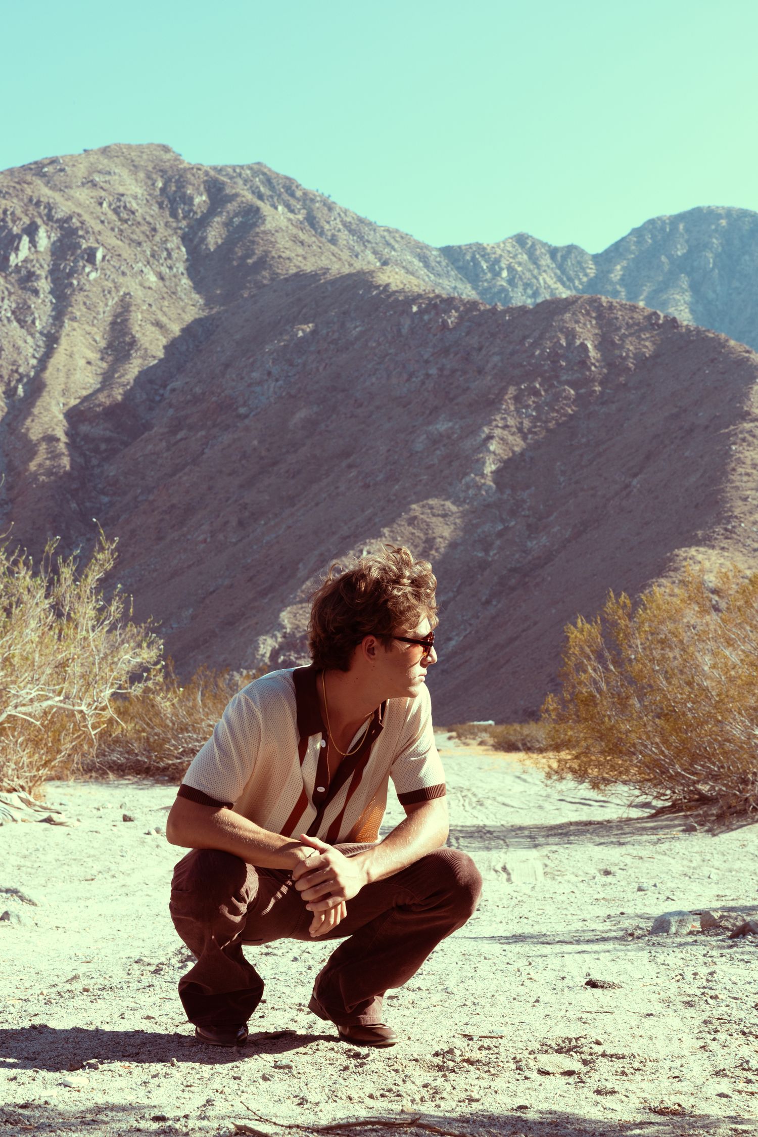 A young man in sunglasses crouches in a desert landscape with rugged mountains and dry shrubs under a clear blue sky.