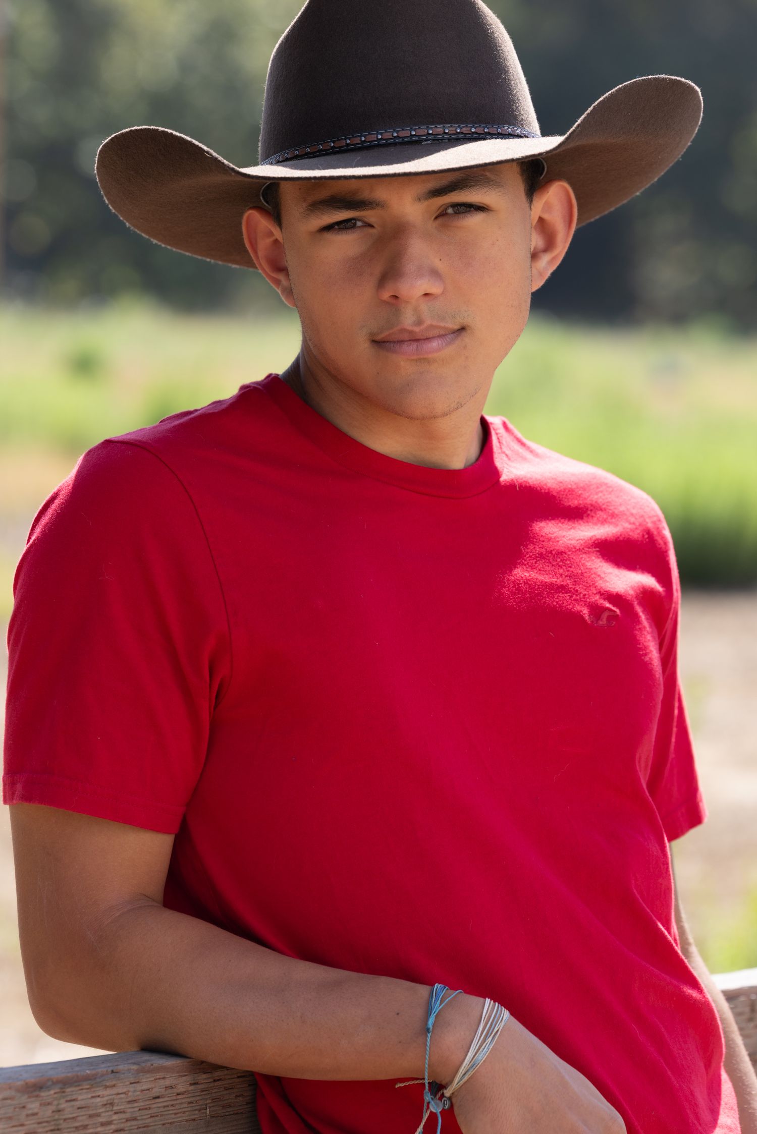 Young man in red t-shirt and brown cowboy hat leaning on a fence outdoors in a sunny rural setting.