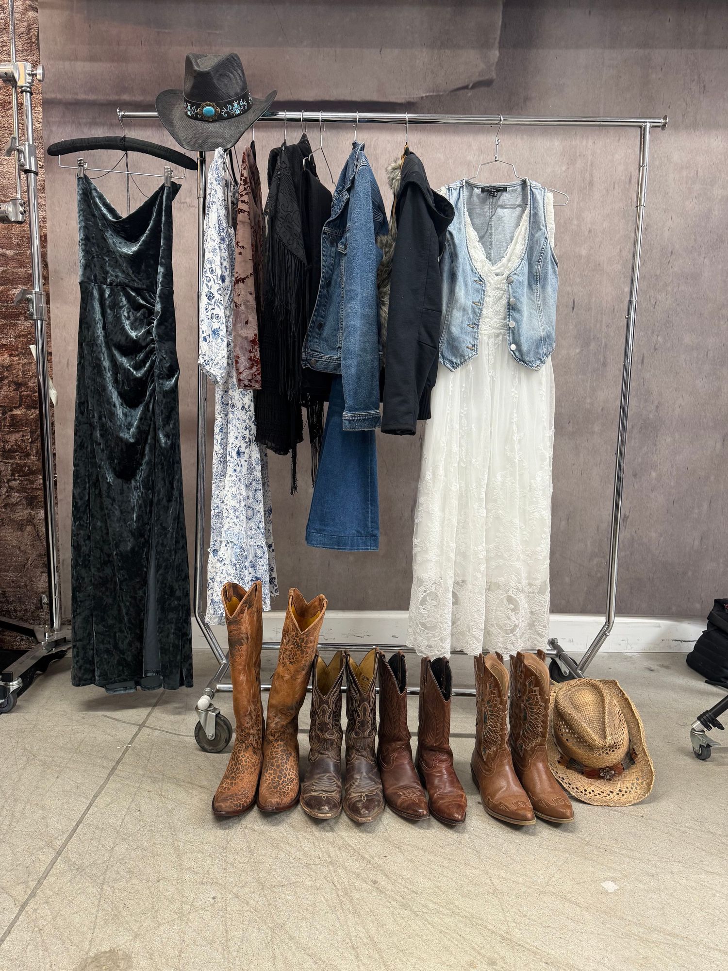 Clothing rack with dresses, denim jacket, cowboy boots, and hats in studio space.