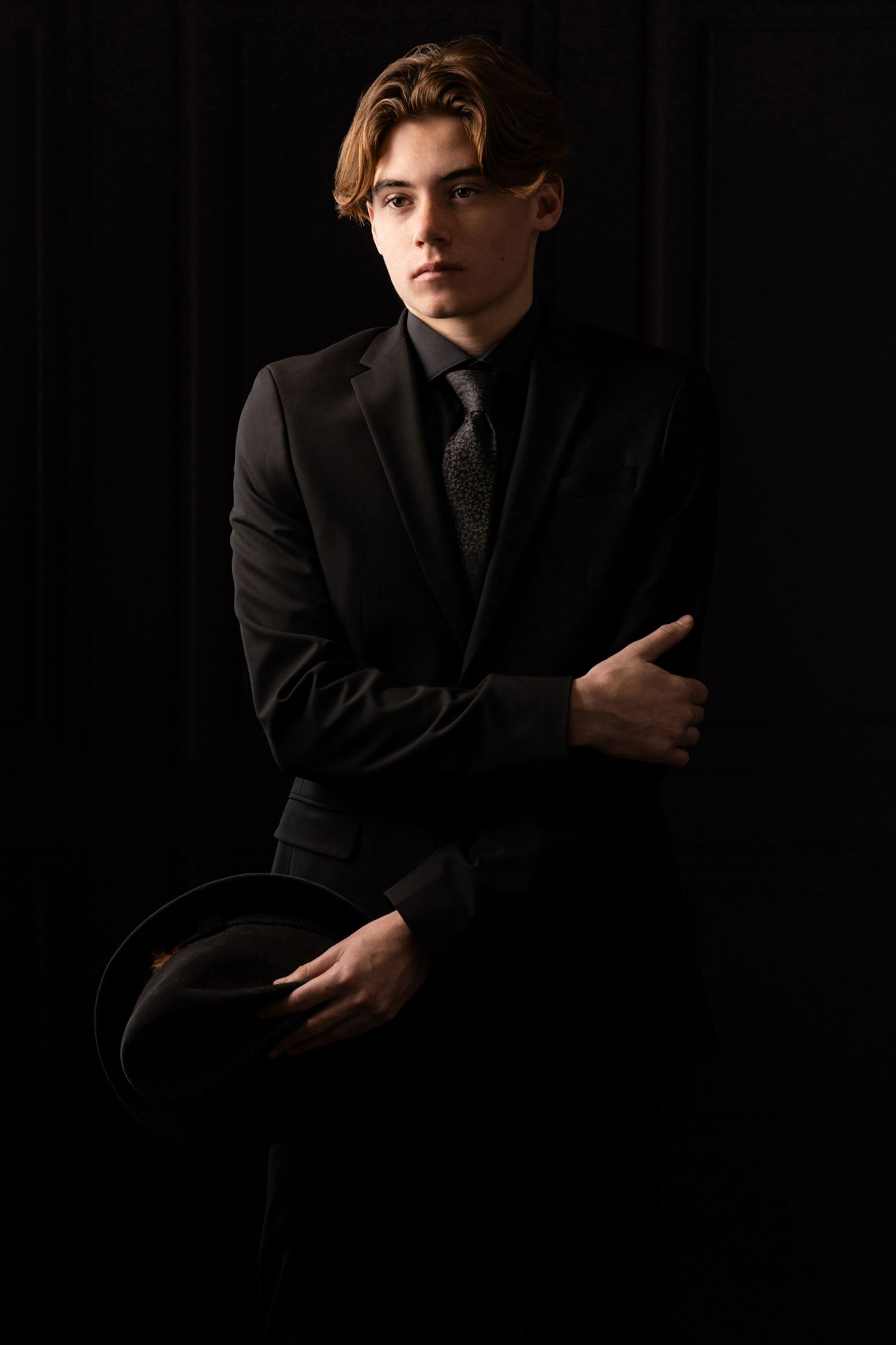 Young man in black suit holding a hat against dark background, elegant formal portrait photography.