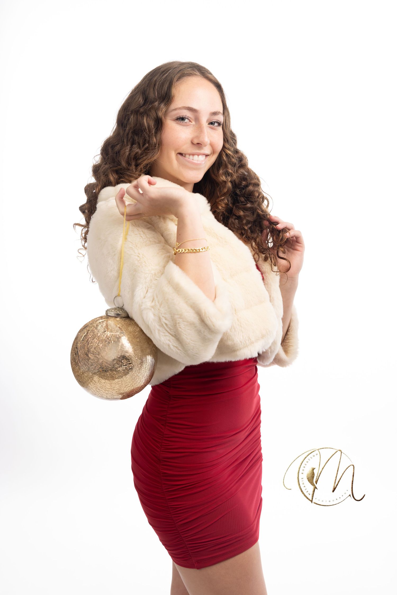 Young woman in red dress and white fur wrap holding gold ornament, smiling over shoulder in holiday portrait.