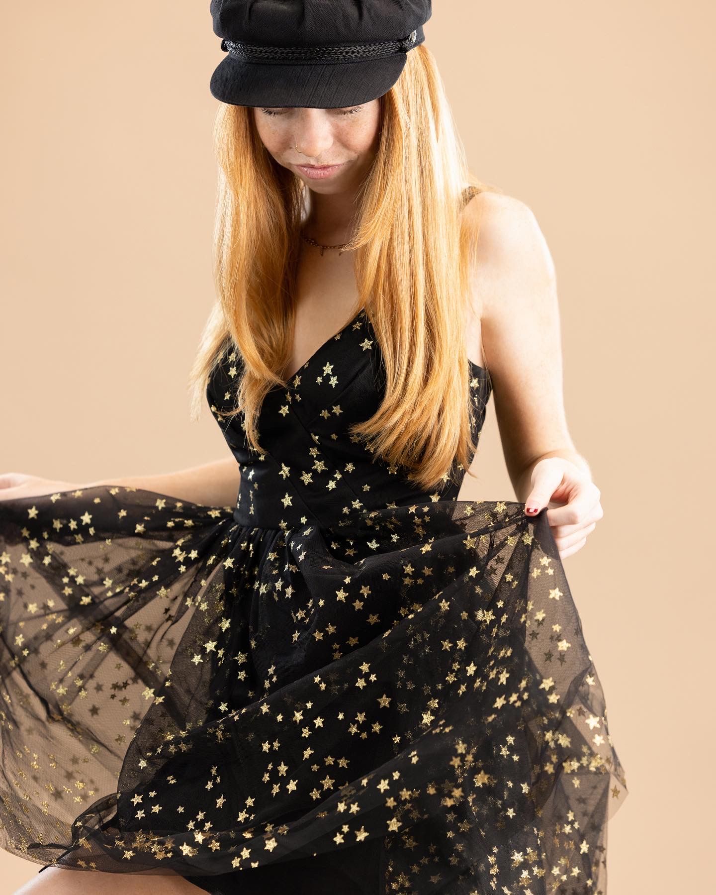 Woman in black star-print tulle dress and baker boy cap, twirling skirt against beige background.