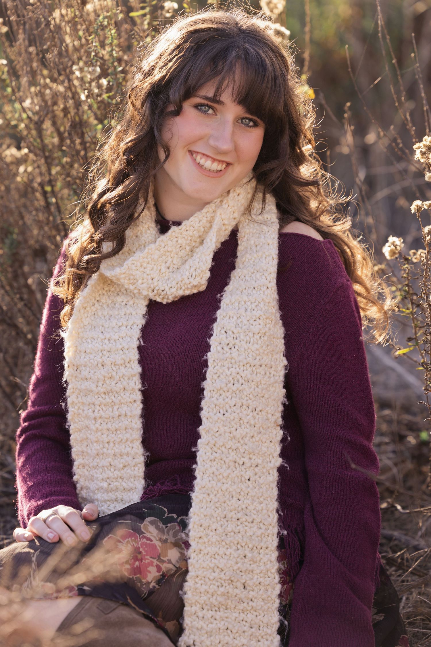 Smiling young woman with curly brown hair wearing a burgundy sweater and cream knit scarf outdoors.