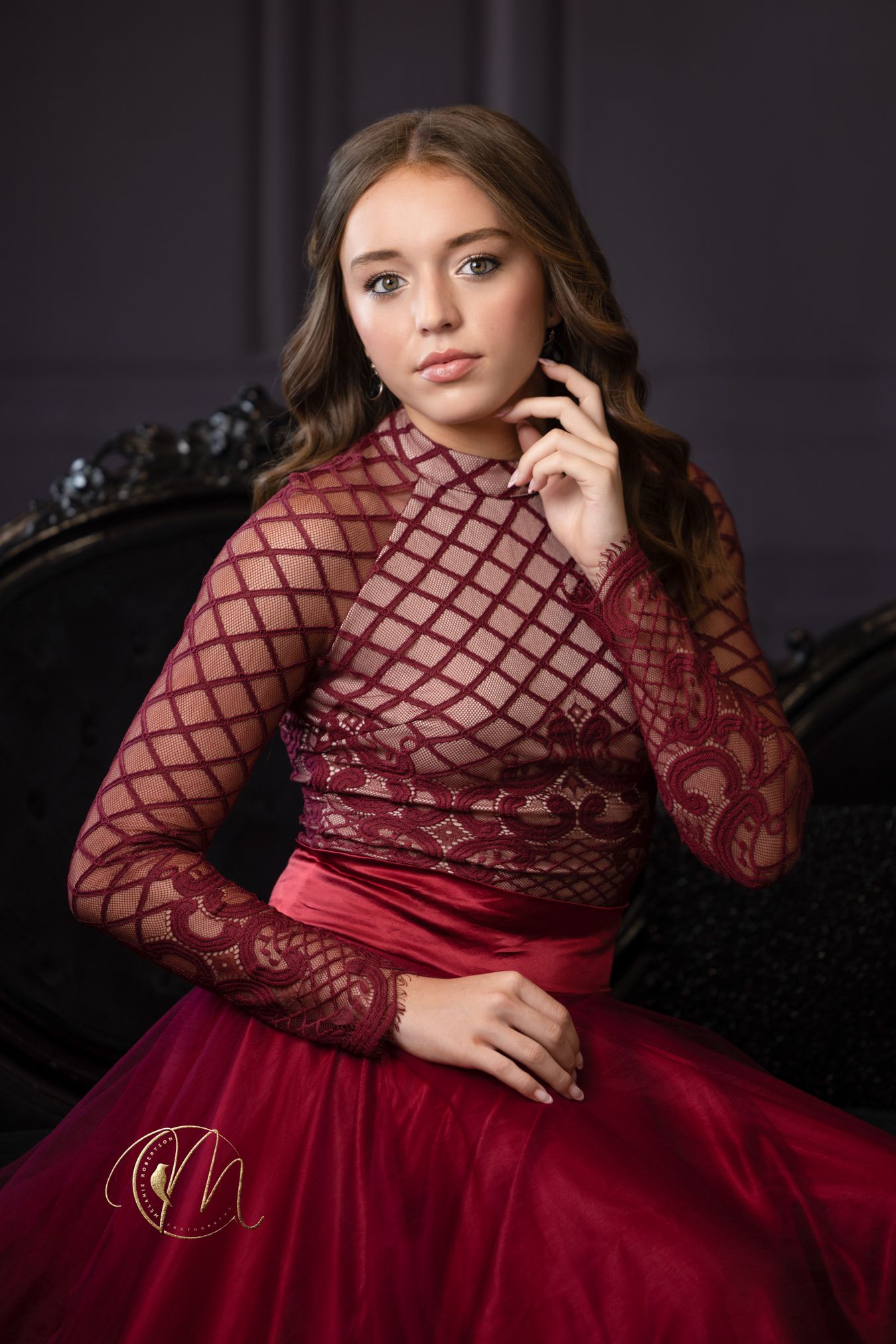 Young woman in elegant burgundy lace gown poses gracefully in a dark studio setting.