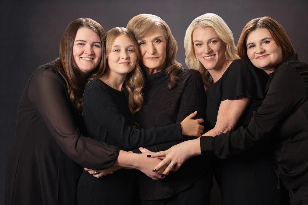 Five women in black outfits pose together in a warm studio portrait, embracing each other against a dark background.