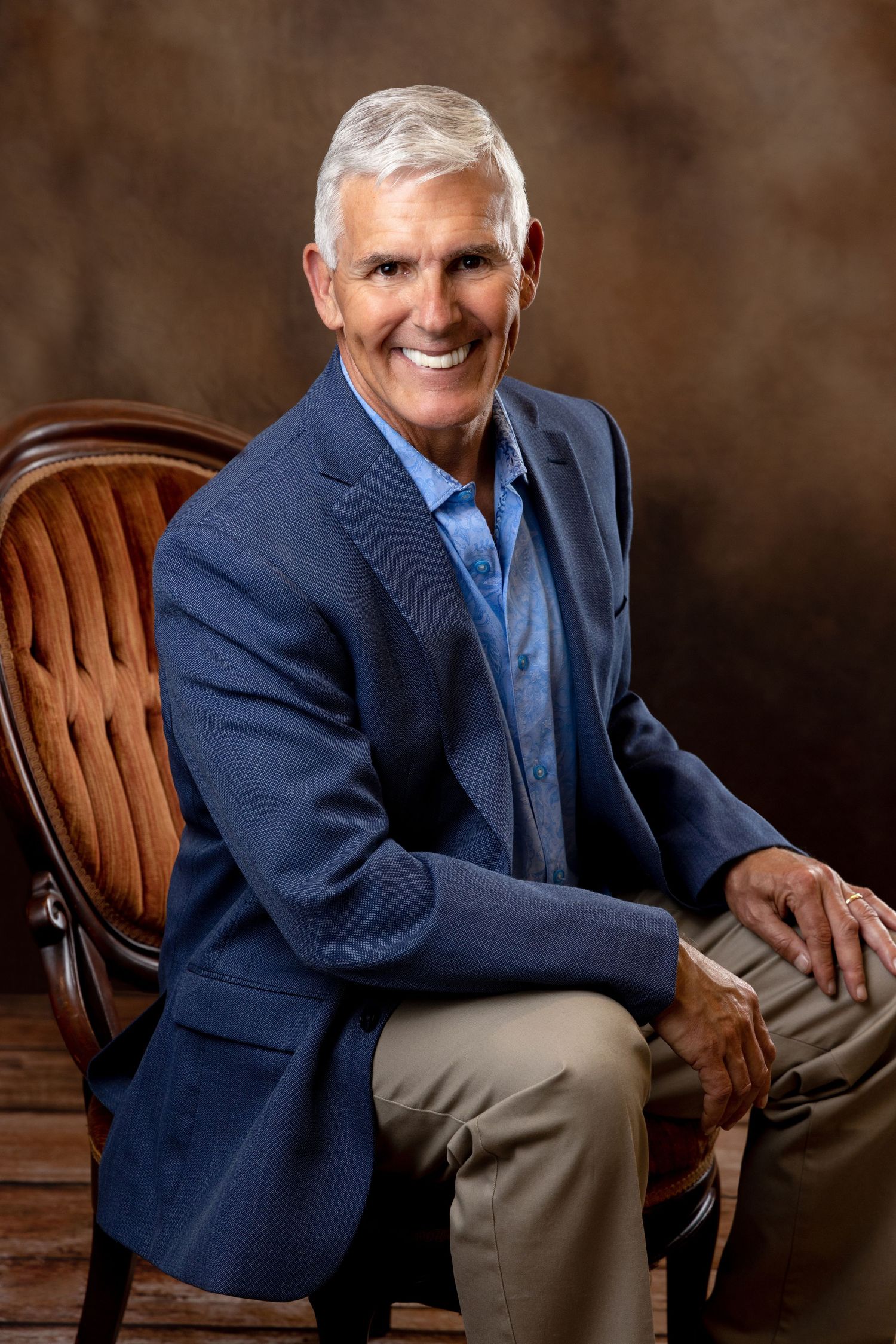 Smiling older man with gray hair sitting in chair wearing blue blazer in professional portrait.