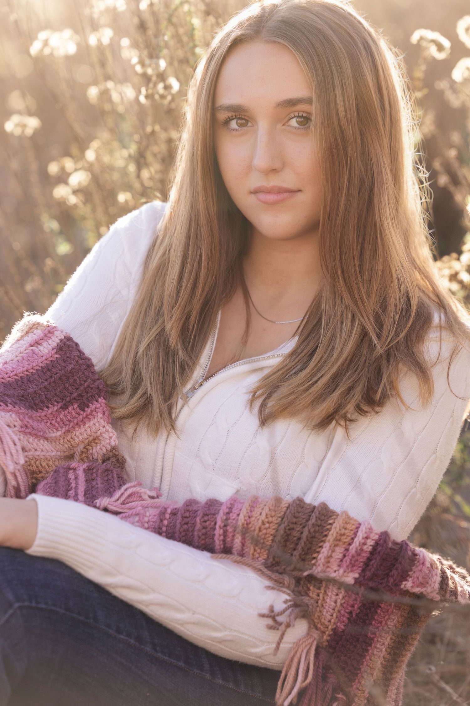 Pretty teen girl in white sweater and pink crochet wrap sits in sunlit golden field.