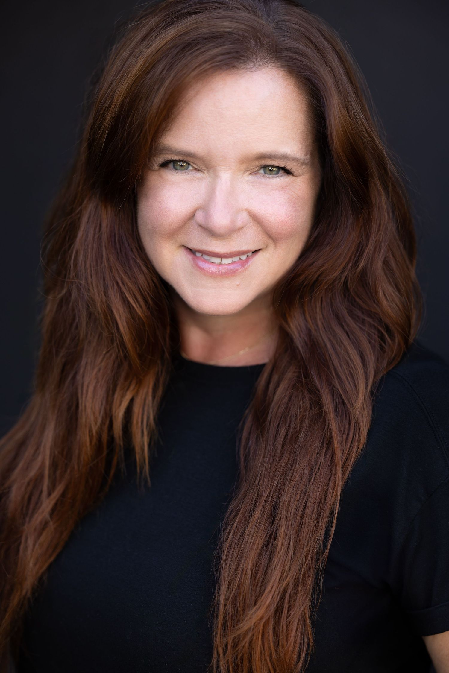 Smiling woman with long auburn hair wearing black top against dark background in professional headshot.
