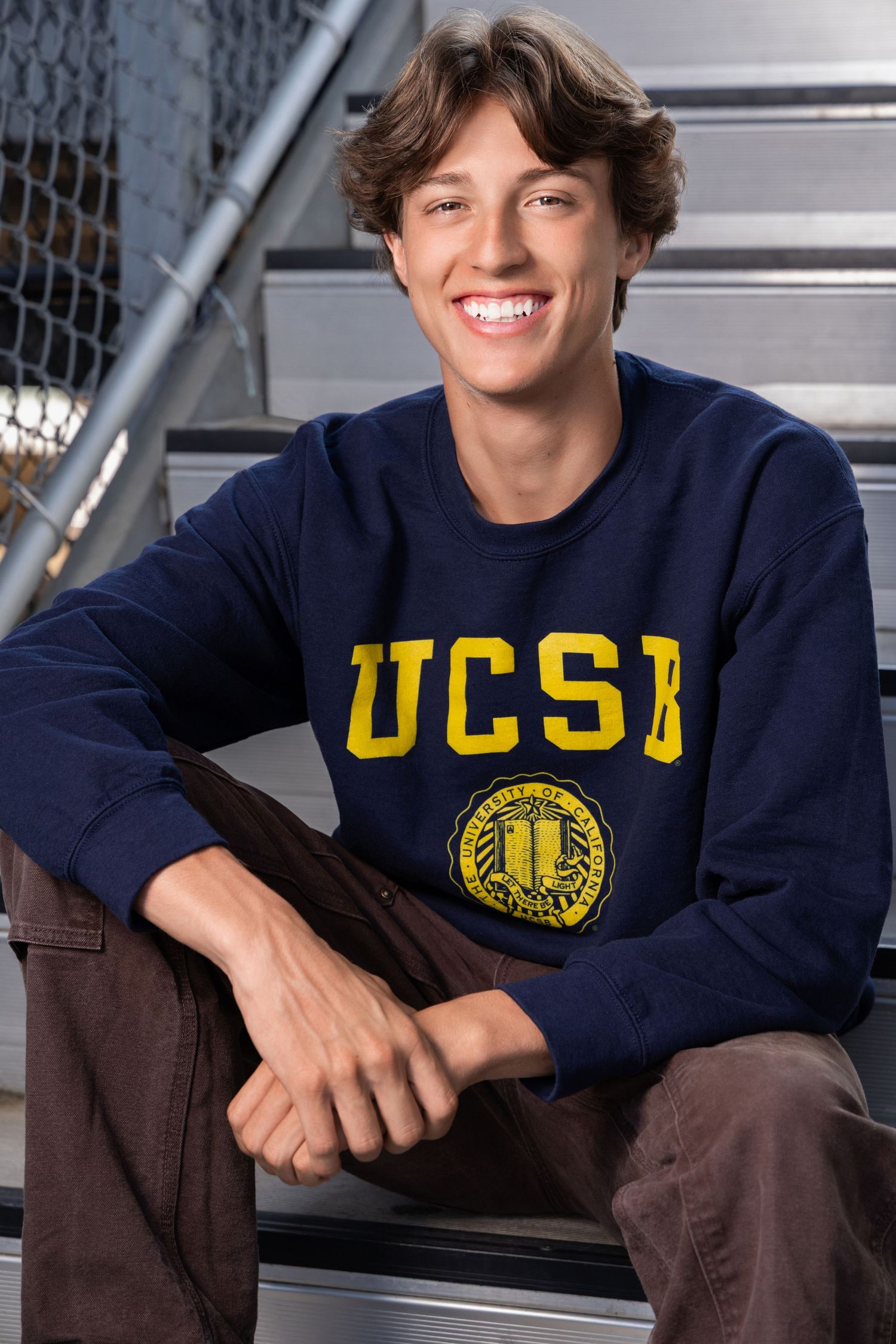 Smiling young man wearing a navy UCSB sweatshirt, sitting on metal bleachers in brown pants.