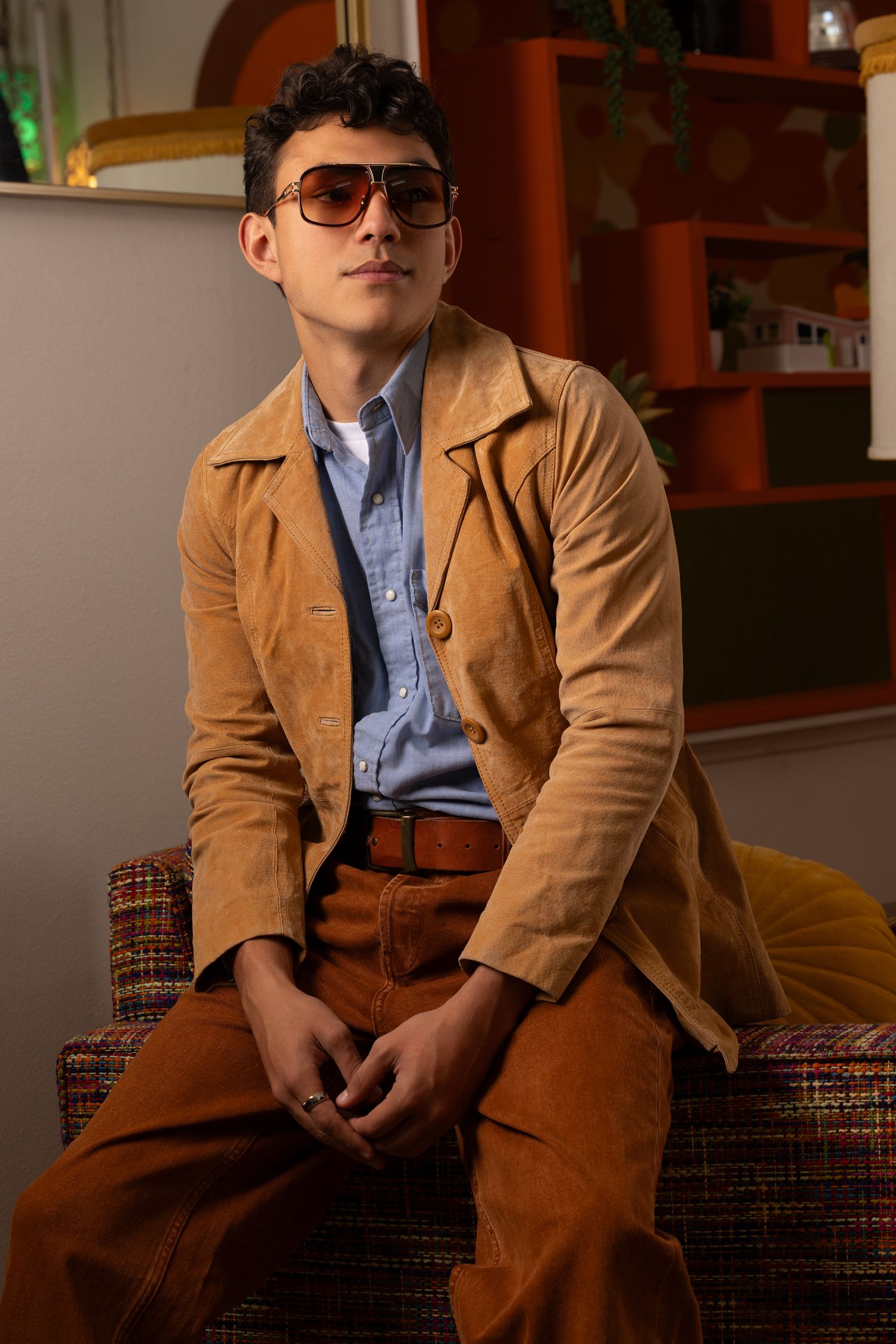 Young man in brown suede jacket, blue shirt, and aviator sunglasses seated in a retro-styled room.
