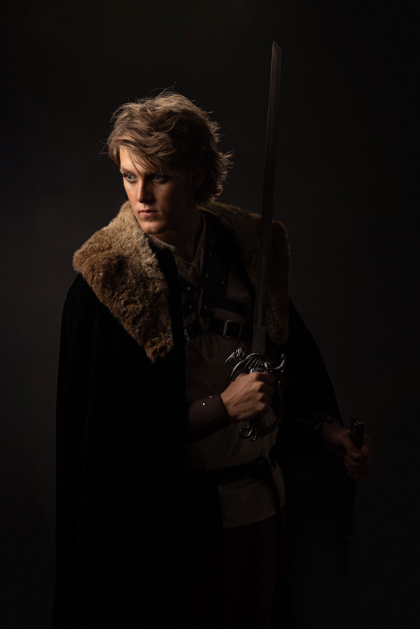 Young medieval warrior in dark cloak with fur collar holding sword against black background.