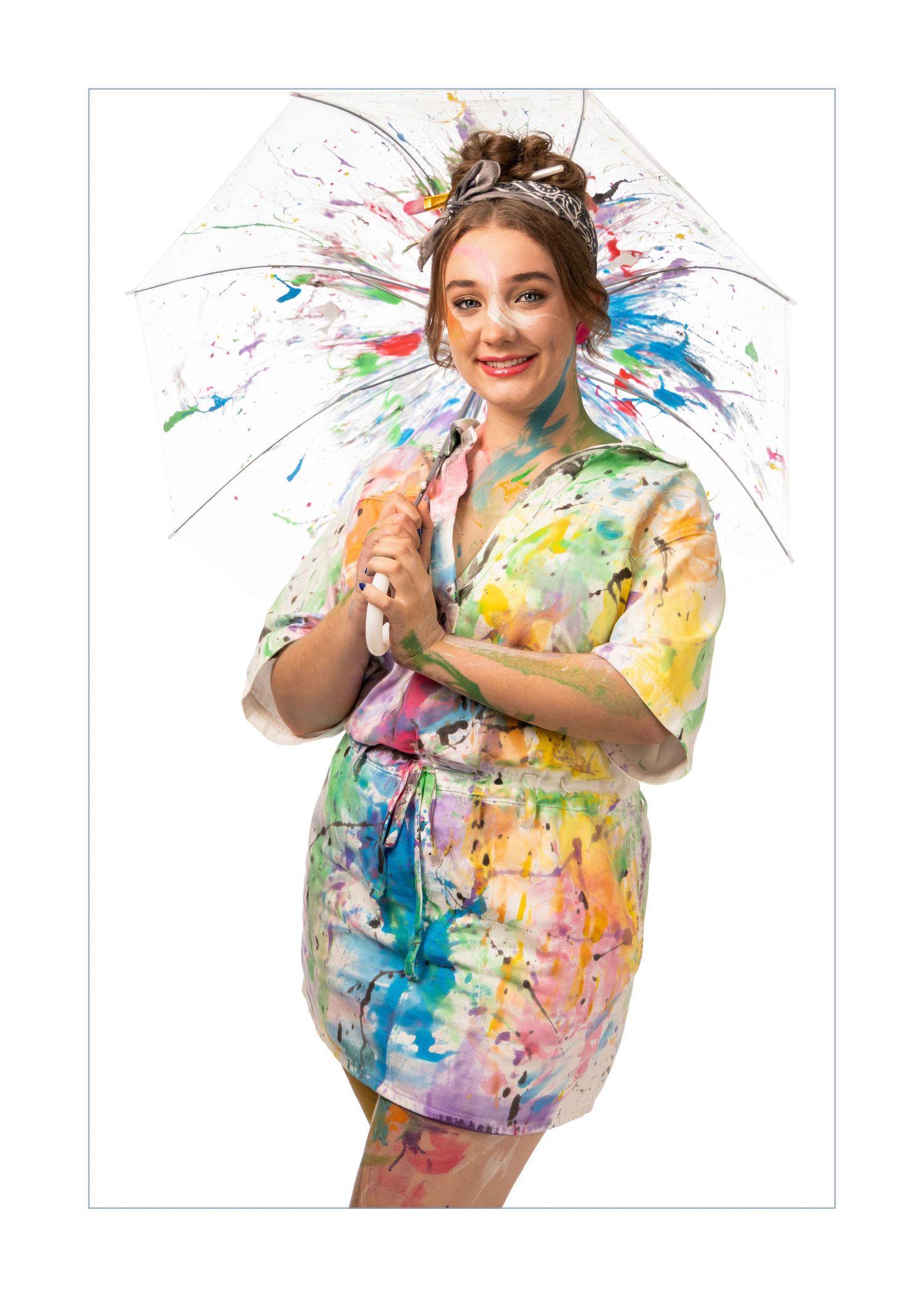 Young woman smiling in colorful paint-splattered outfit holding clear umbrella with vibrant paint splashes.