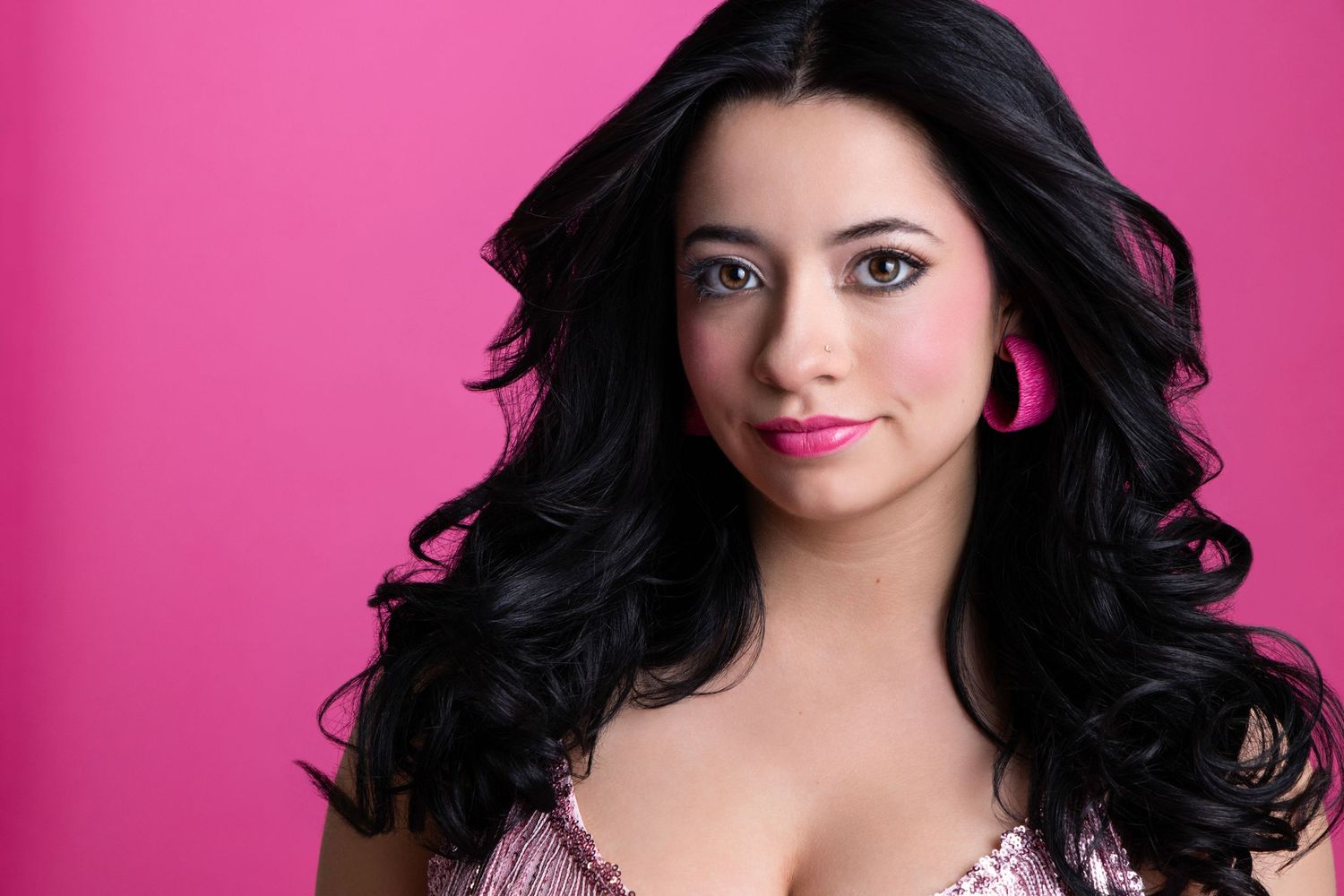 Young woman with long dark wavy hair poses against a vibrant pink background wearing pink earrings and makeup.
