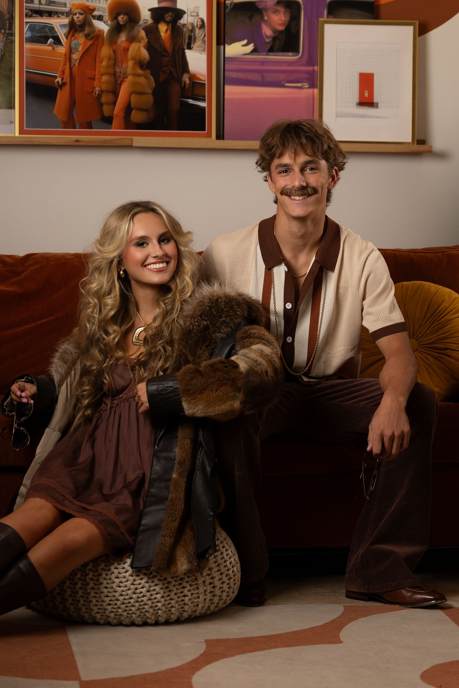 A smiling couple in 1970s retro outfits sit on a brown velvet sofa with vintage-style artwork on the wall behind them.