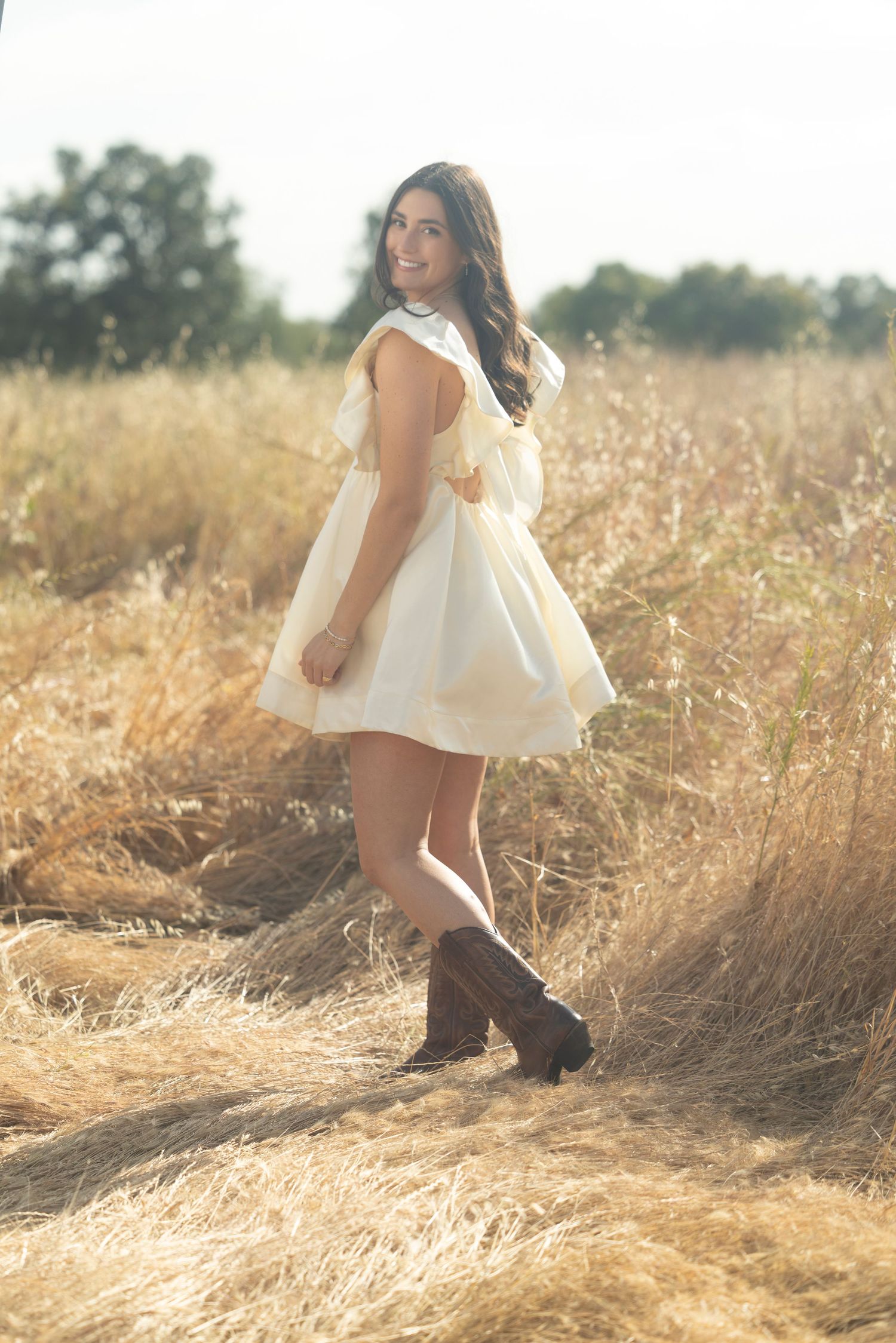 Smiling woman in a flowy cream mini dress and brown cowboy boots walks through a golden dry grass field under soft natural light.