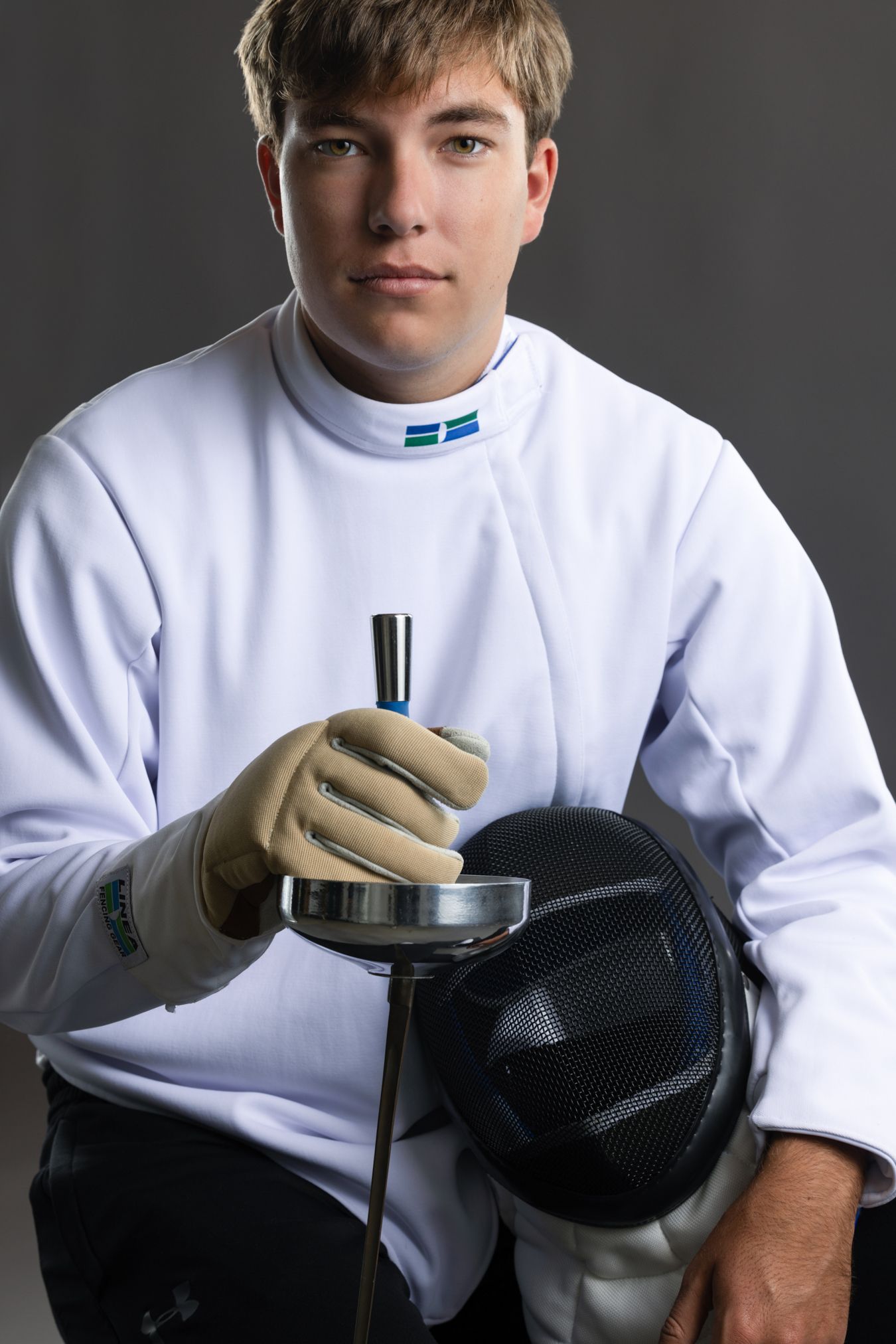Young male fencer in white uniform holding épée and mask in studio portrait.