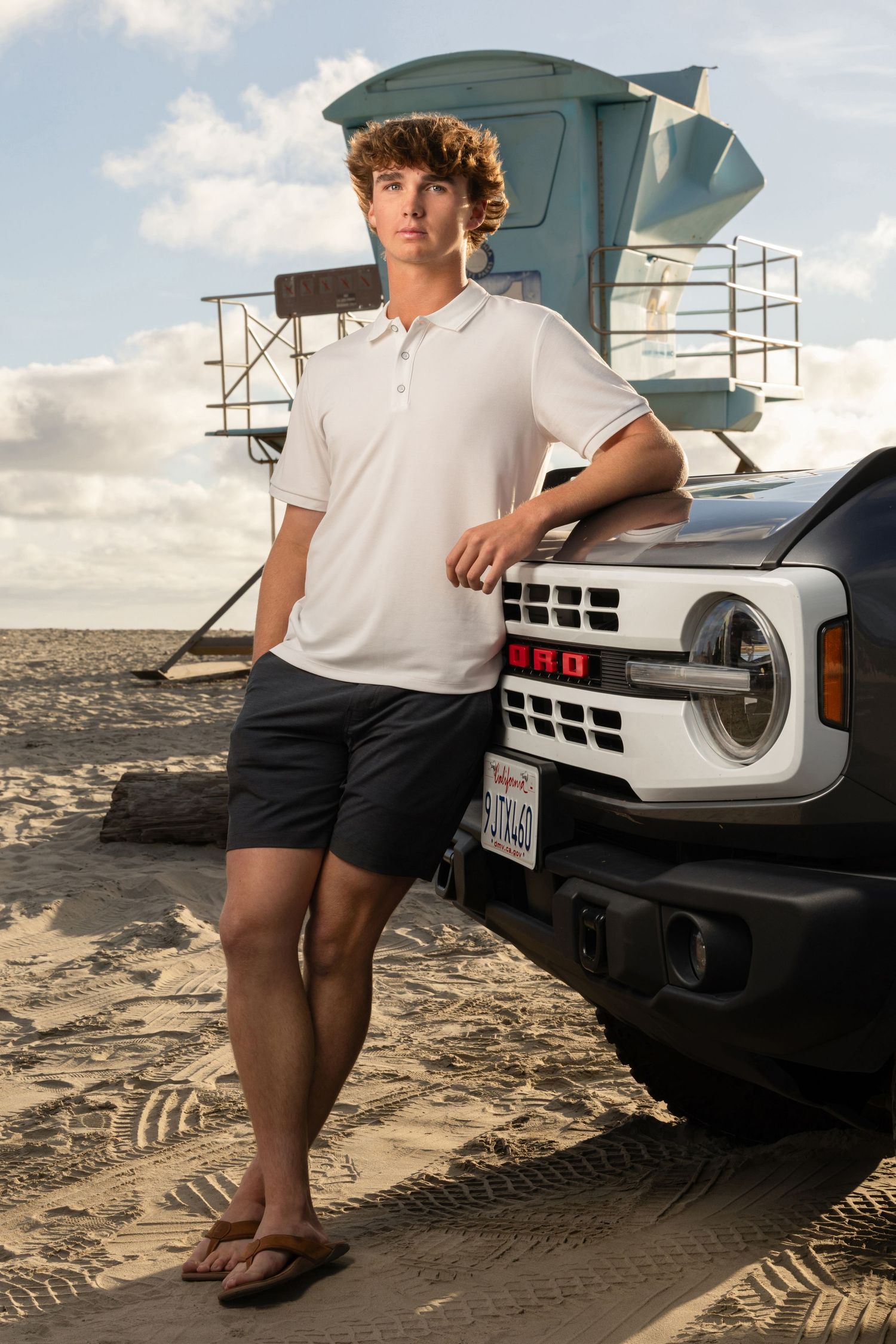 Teen boy in white polo and shorts leans on a Ford Bronco on the beach near a lifeguard tower.