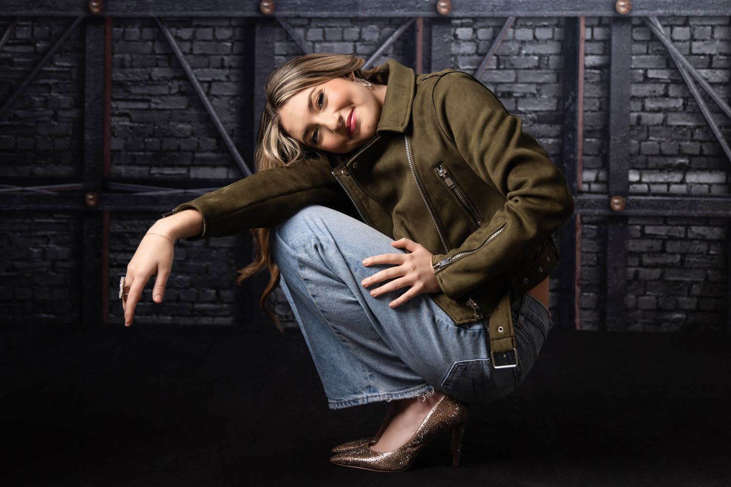 Confident blonde woman in olive jacket and jeans posing against dark brick wall background.