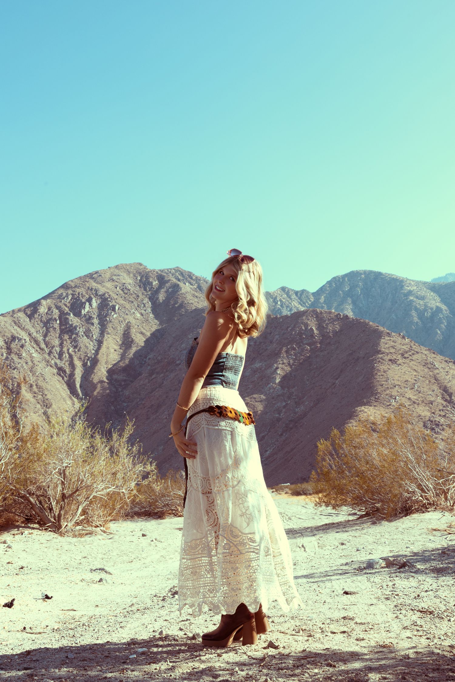 Blonde woman in white lace maxi dress and boots posing in desert landscape with mountains and clear blue sky.