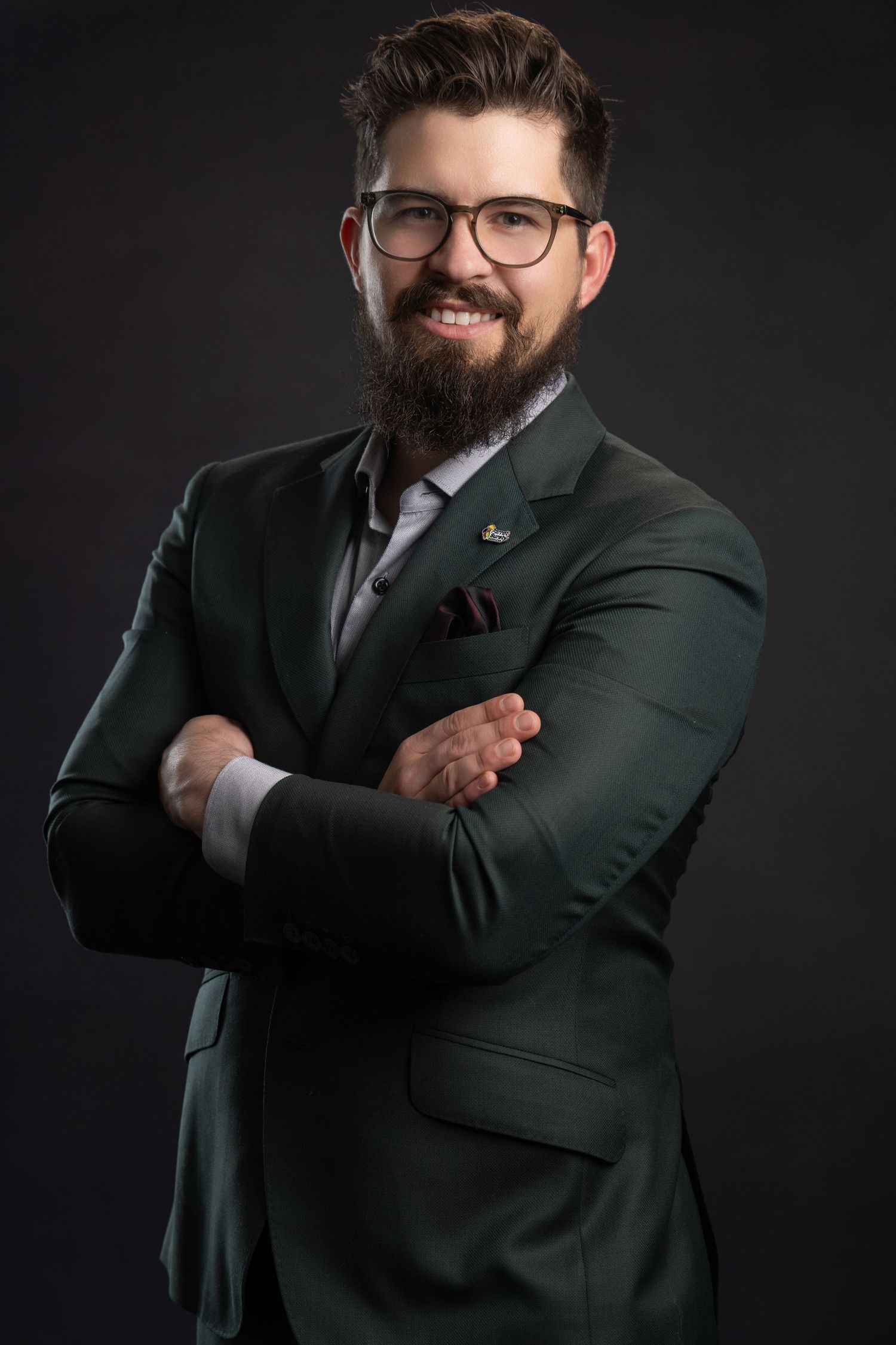 Smiling bearded man in green suit with glasses, arms crossed, professional studio portrait on dark background.