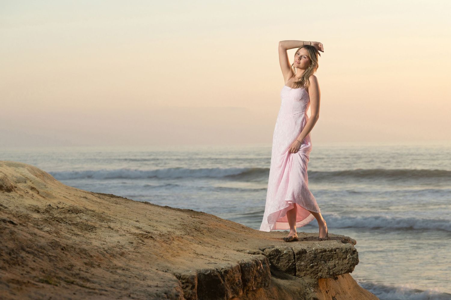 Woman in pink dress posing on coastal rocks at sunset with ocean waves in background.
