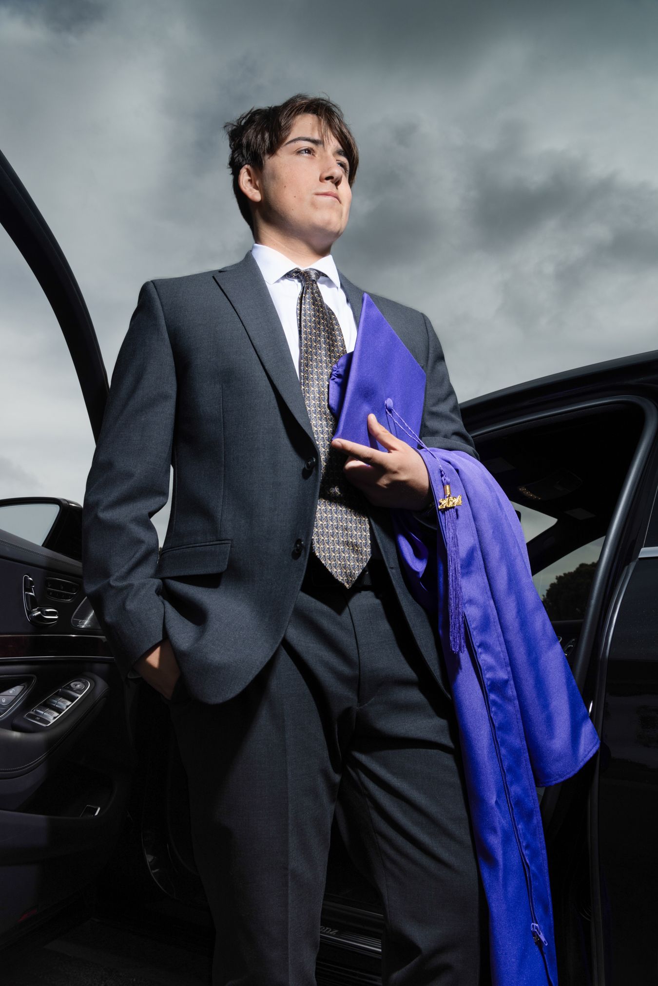 Young man in gray suit holding blue graduation gown, standing beside luxury car under dramatic cloudy sky.