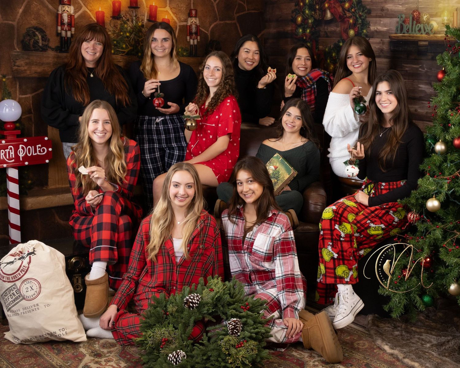 Group of women in festive plaid pajamas posing together near a Christmas tree and fireplace for a holiday photo.