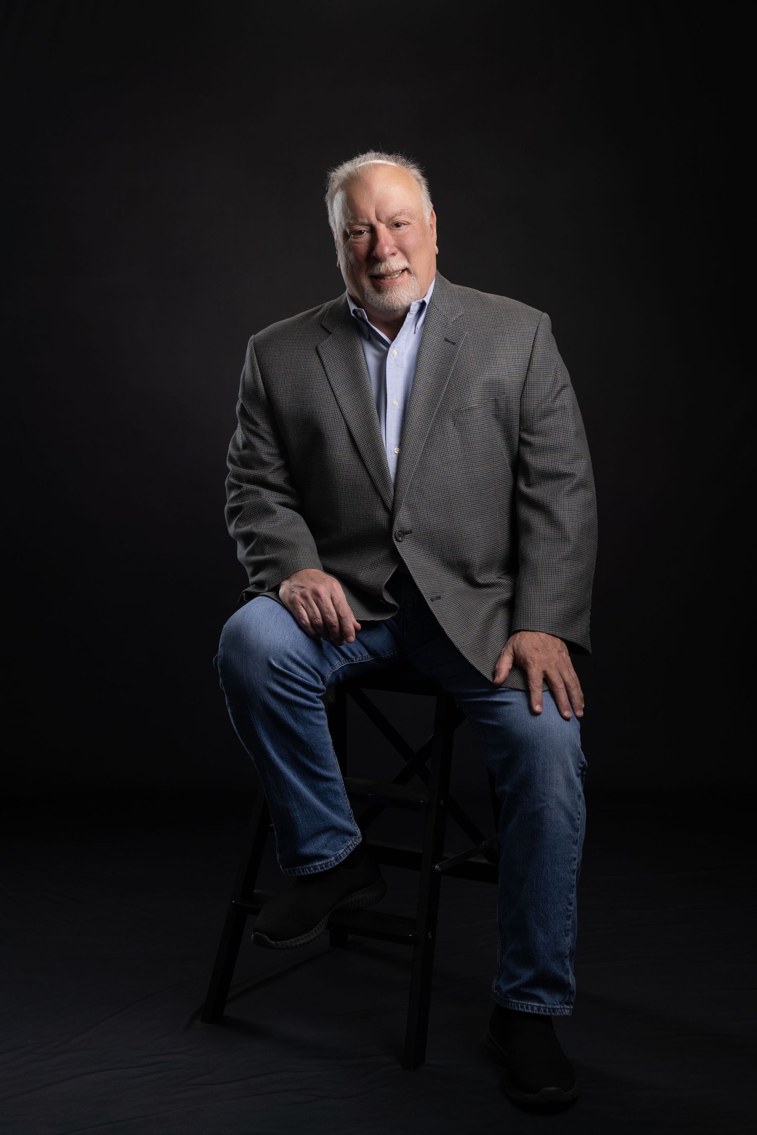 Older man with white hair in gray blazer and jeans sits on stool against dark background, smiling warmly.