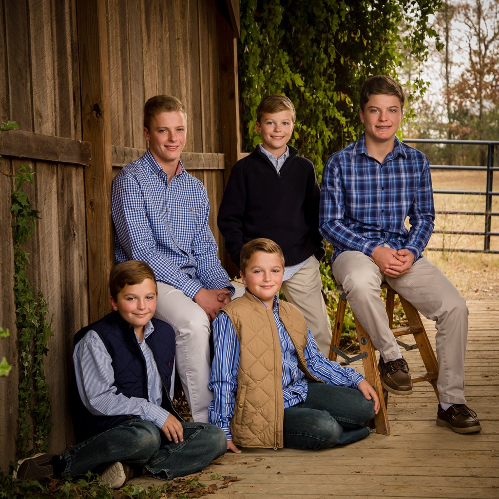 Four boys in casual preppy attire posing for outdoor family portrait near rustic wooden barn.
