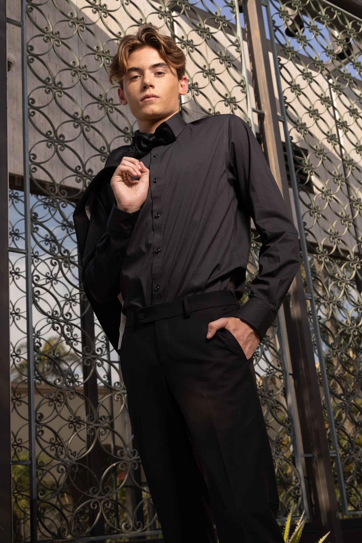 A young man in a black dress shirt and trousers poses against an ornate iron gate outdoors.
