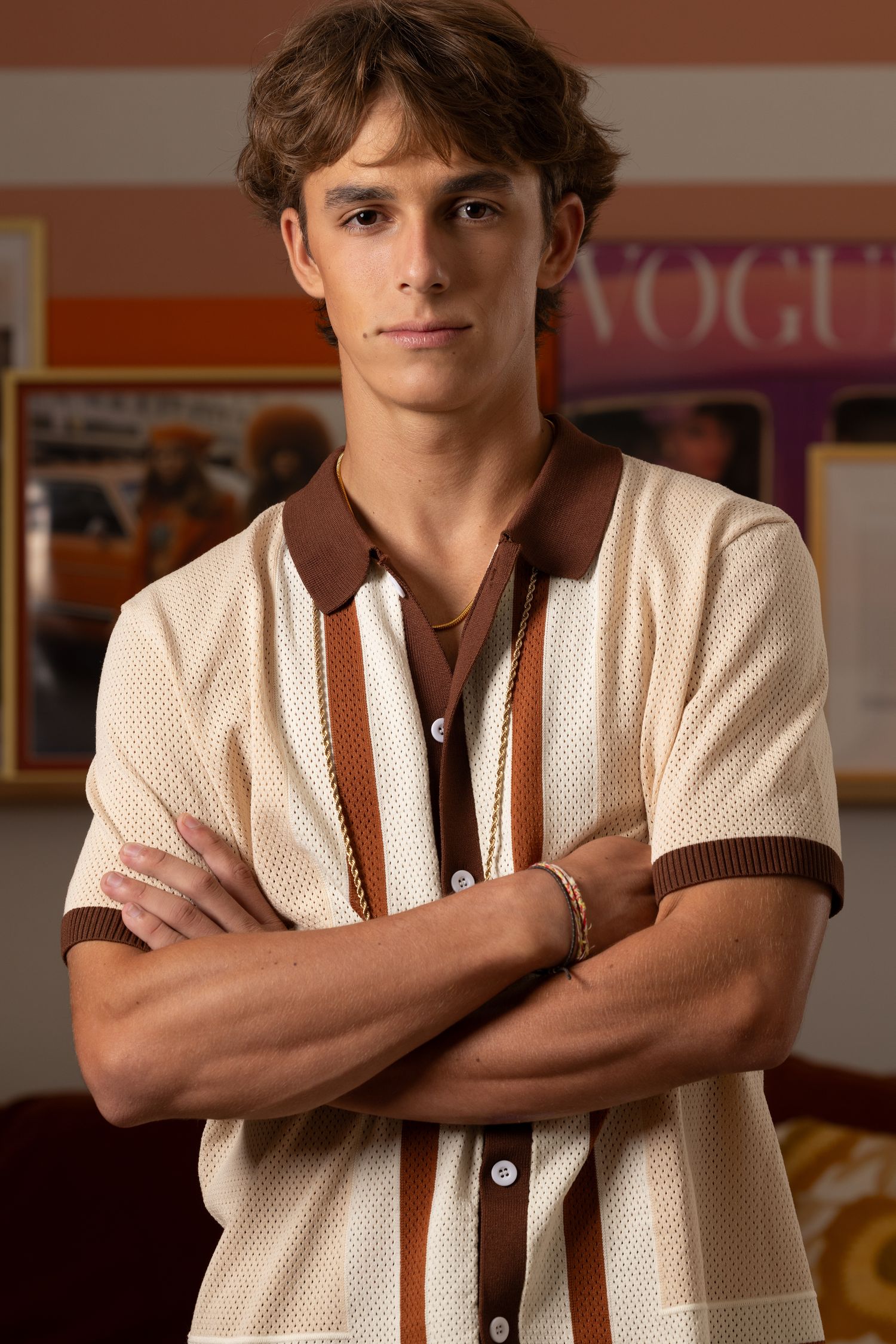 Young man with brown hair, arms crossed, wearing a retro cream and brown polo shirt, standing indoors.