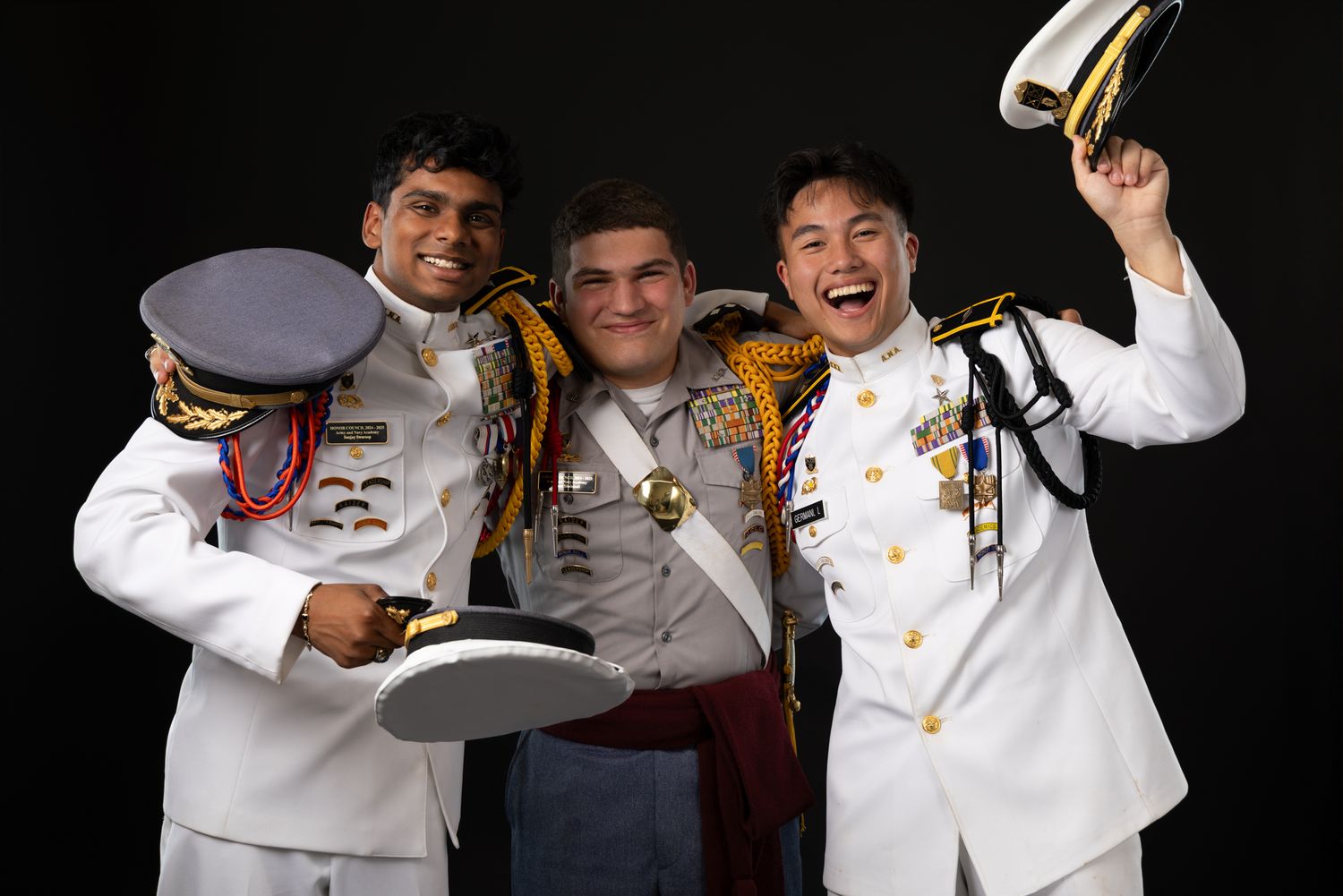 Three young men in military dress uniforms smile and celebrate together against a dark studio background.