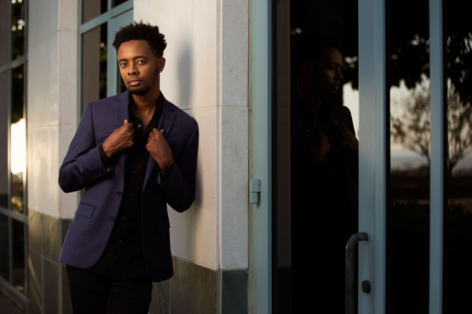 Stylish young man in a navy blazer leaning against a glass door at dusk outdoors.