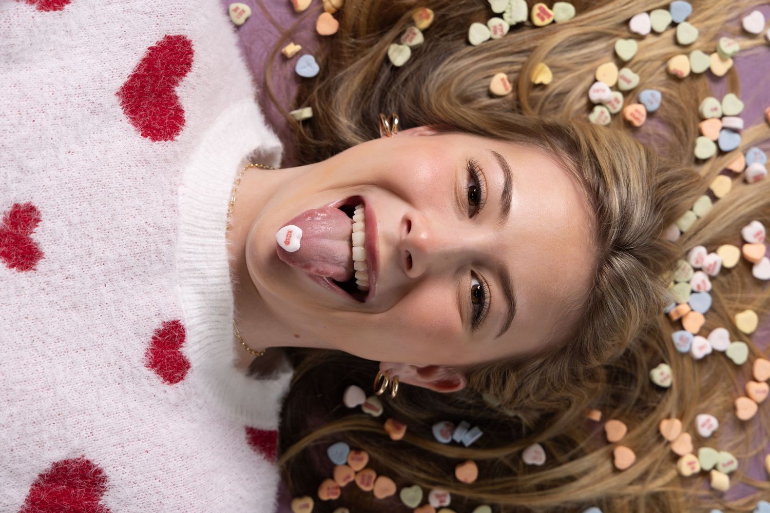 Young woman lying down smiling with candy hearts in her hair, wearing a heart-print sweater on Valentine's Day.