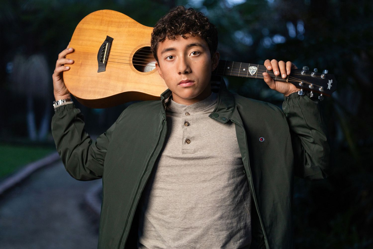 A young man holds an acoustic guitar over his shoulder in an outdoor evening portrait setting.