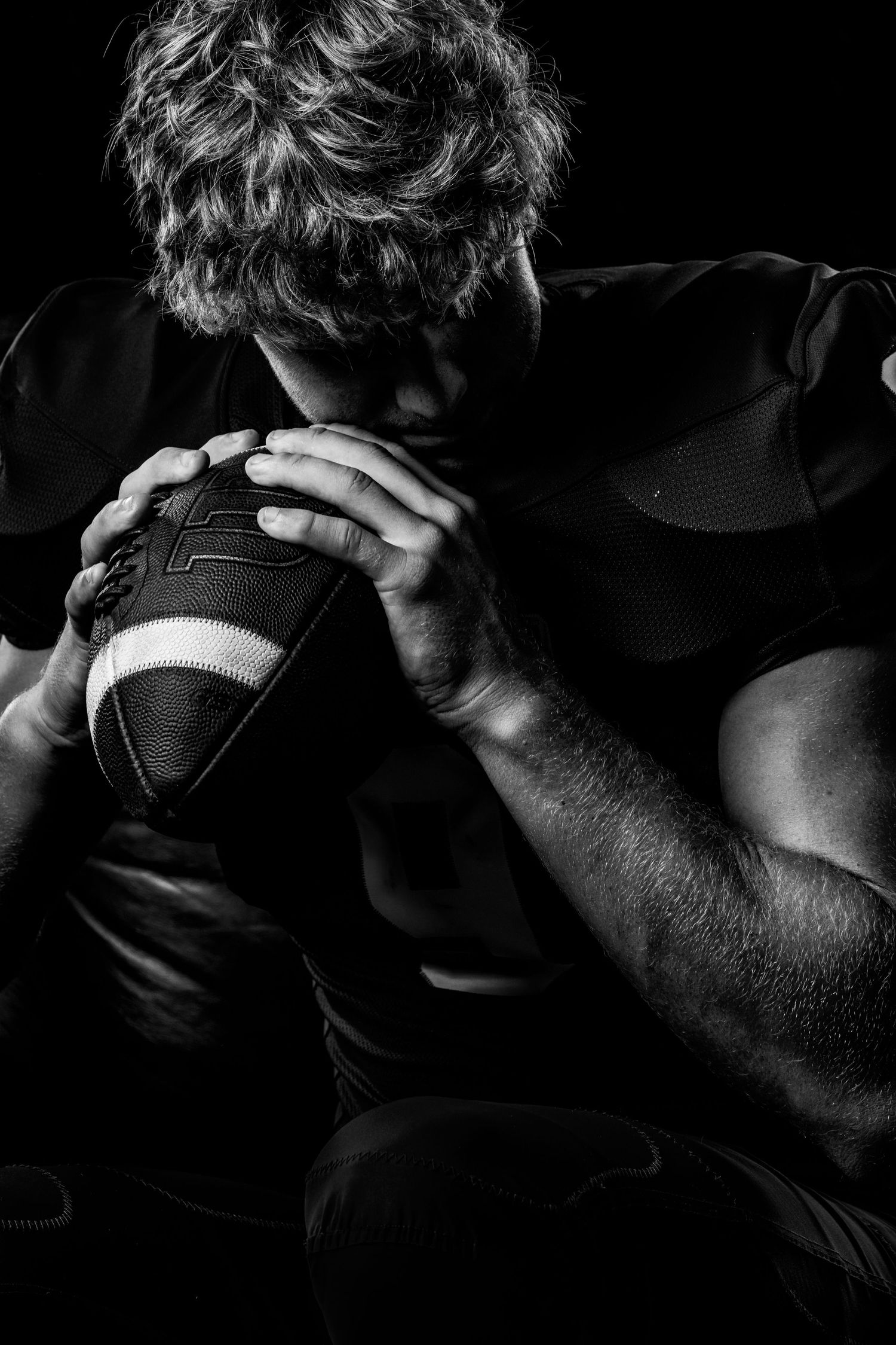 Black and white photo of a muscular American football player holding a football close to his face in dramatic lighting.