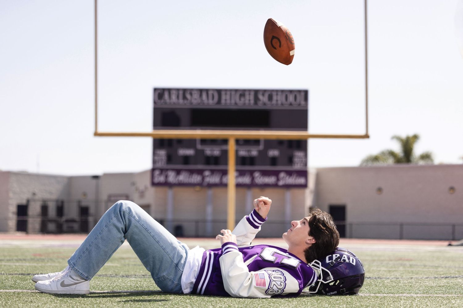 Teen athlete lying on football field tossing ball at Carlsbad High School stadium.