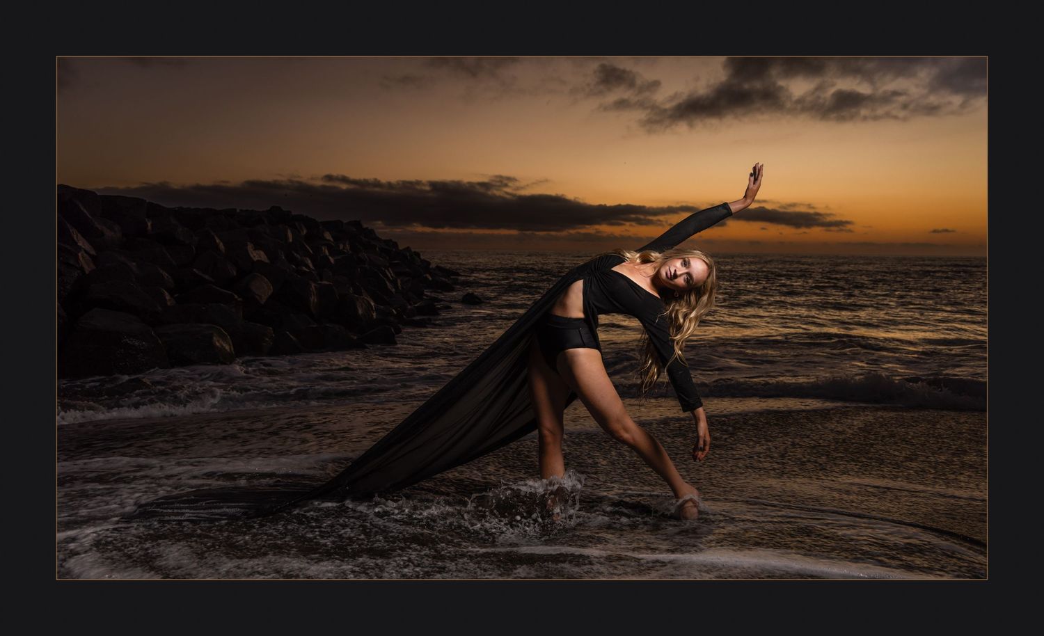 Dancer poses dramatically on beach at sunset wearing flowing black fabric.