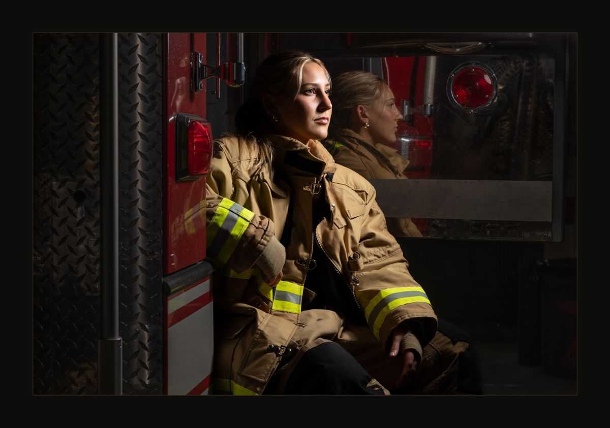Female firefighter in turnout gear sitting pensively beside a red fire truck in dramatic lighting.