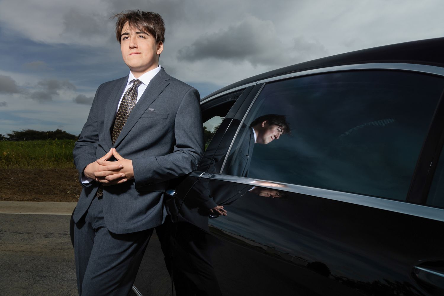 Professional man in gray suit leaning against a black luxury car outdoors under dramatic cloudy sky.