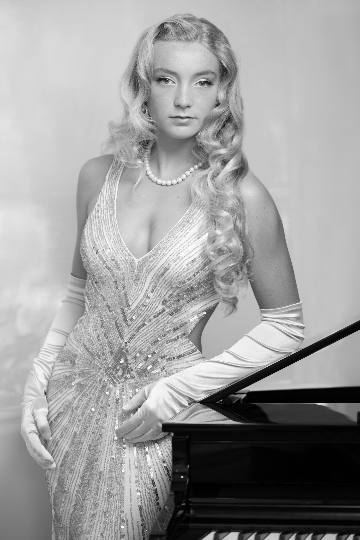 Elegant blonde woman in beige sequin gown and white gloves posing by a grand piano in black and white.