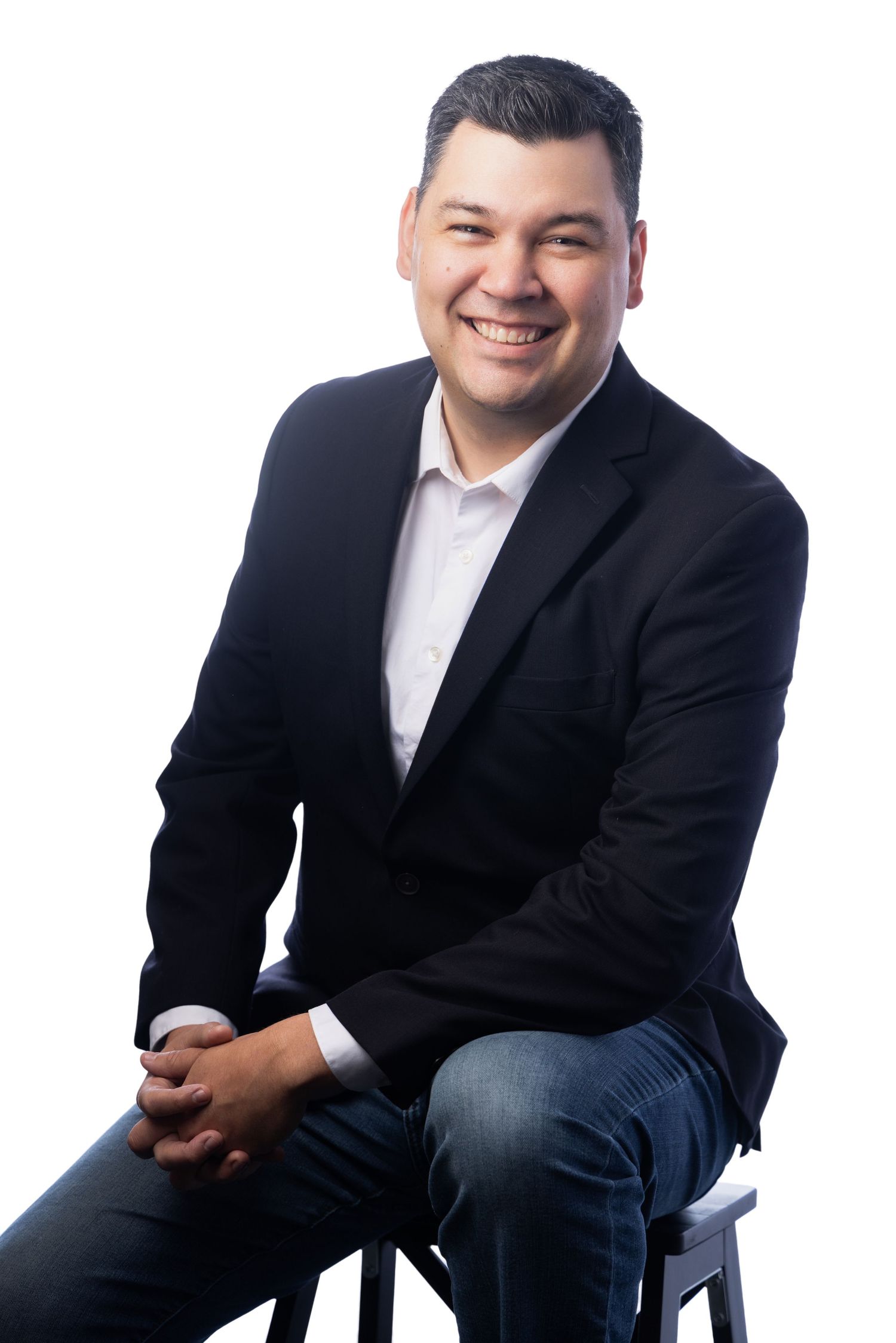 Smiling man in black blazer and jeans sits confidently on a stool against a white background.