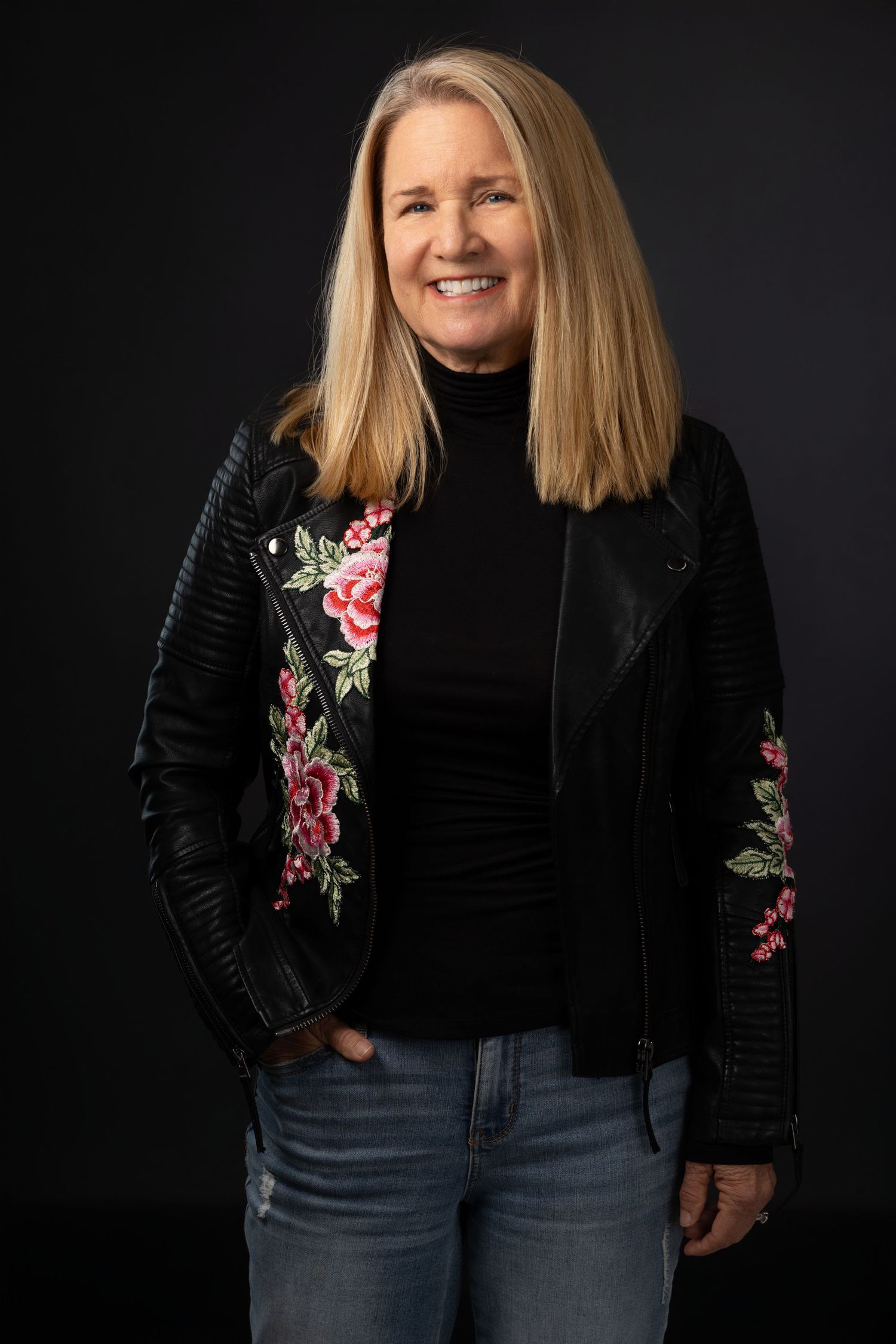 Smiling woman with blonde hair wearing a black floral embroidered leather jacket and jeans against dark background.