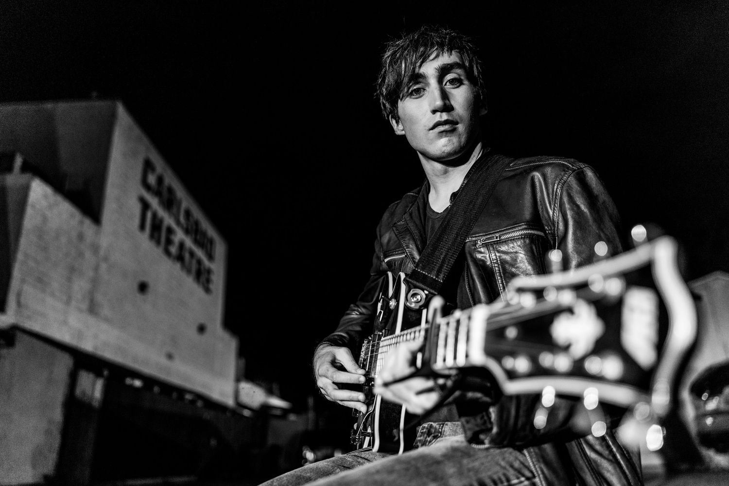 Young guitarist in leather jacket poses outdoors at night near Carlson Theatre in black and white photo.