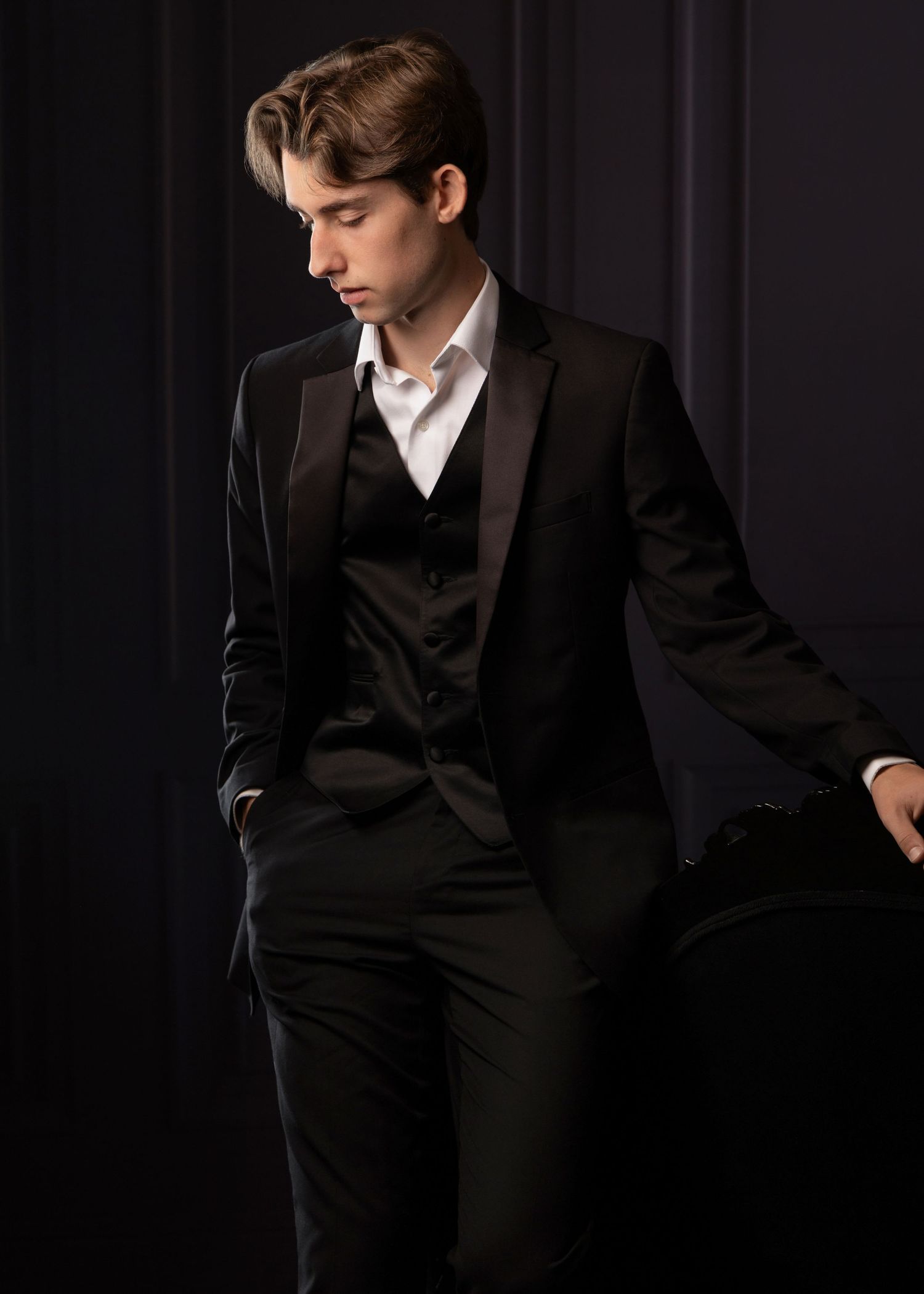 Young man in black three-piece tuxedo suit posing against dark background, looking downward with hands in pockets.