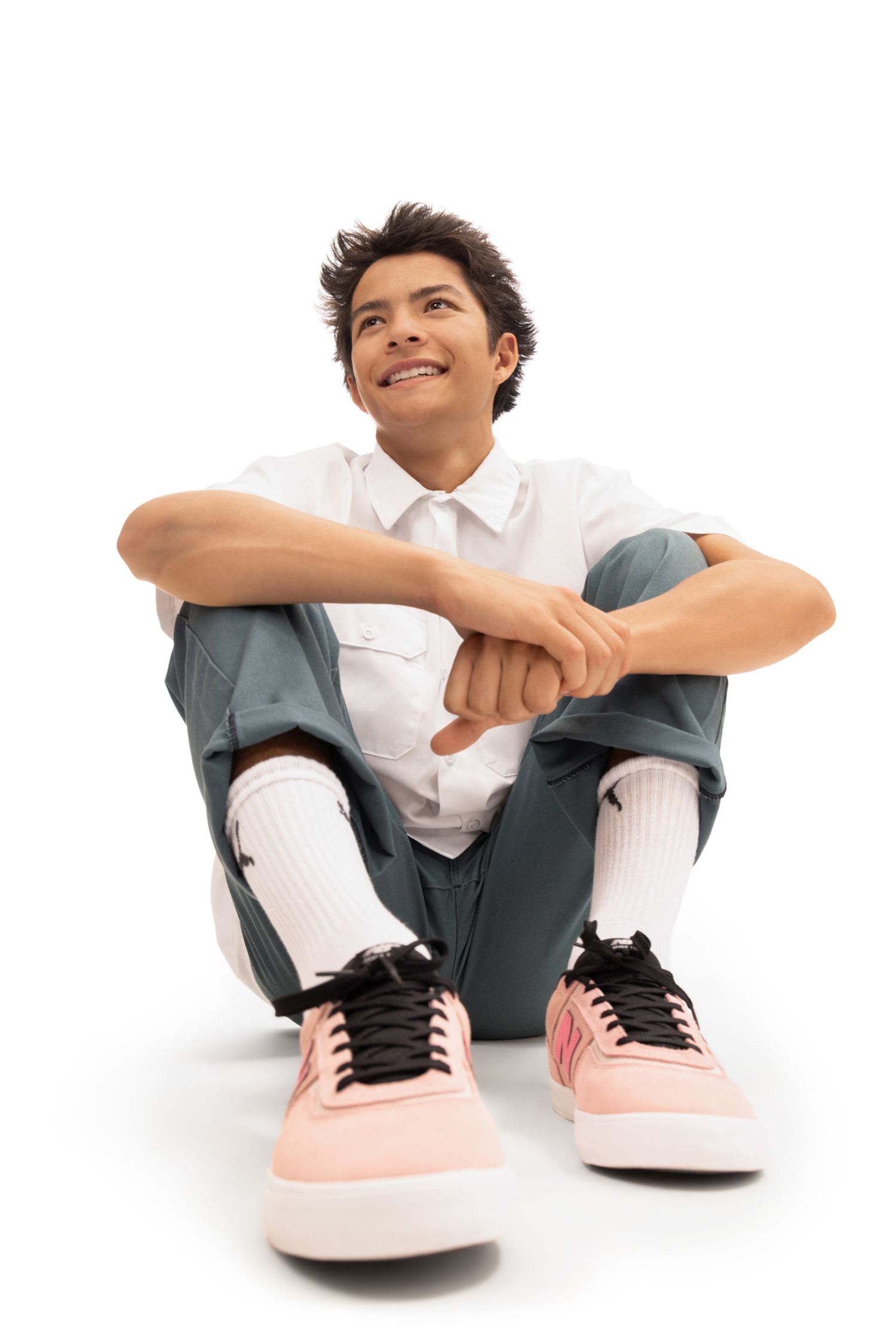 Young man sitting on floor in white shirt, gray pants, white socks, and pink sneakers, smiling and looking up.